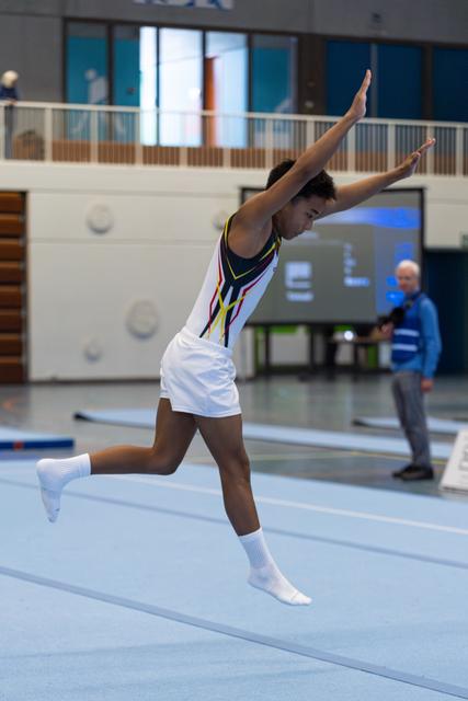 Athlete performs graceful floor leap with arms raised, one leg bent, white socks on blue mat in indoor training facility