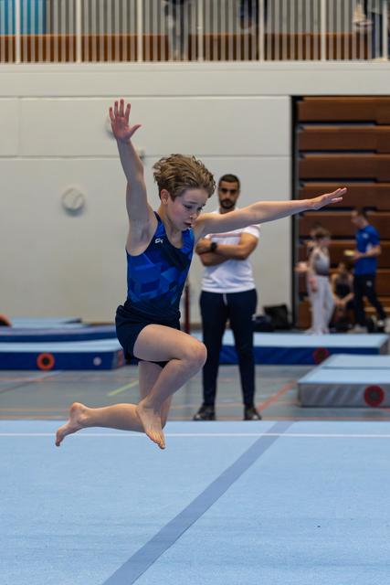 Young athlete performs a graceful leap on floor exercise, arms extended, with coach observing in background
