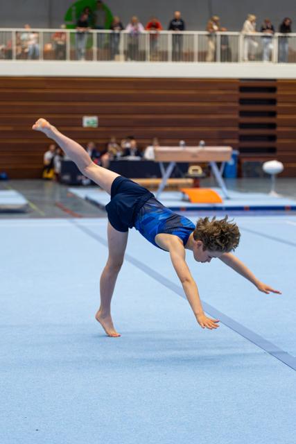 Young athlete performs a one-handed bridge position on floor exercise during indoor meet with spectators watching above