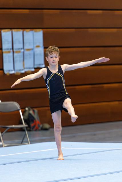 Young gymnast performs a balanced standing pose with arms extended during floor routine, displaying focus and control