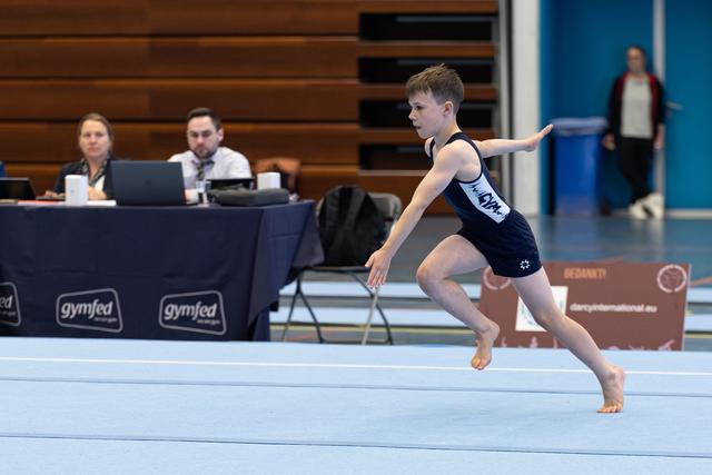 Young athlete performs floor routine with focused expression, arms extended, as judges watch from table in background