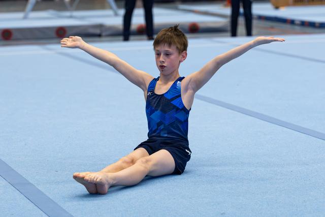 Young athlete performs floor routine in seated position with arms extended, displaying focus and form on blue mat