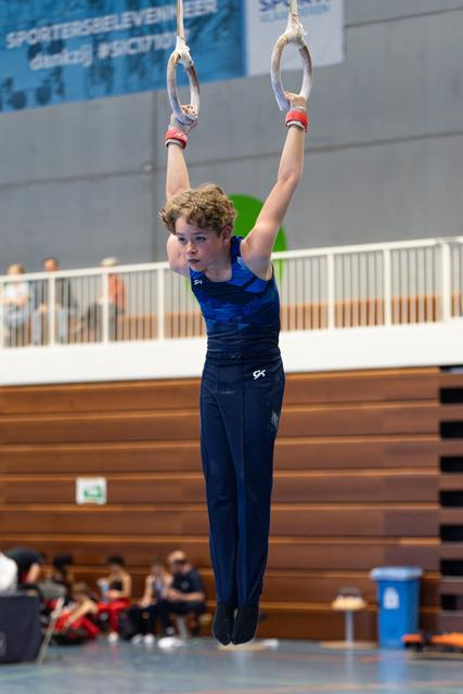 Young athlete demonstrates strength on still rings with focused expression during routine at indoor sports hall