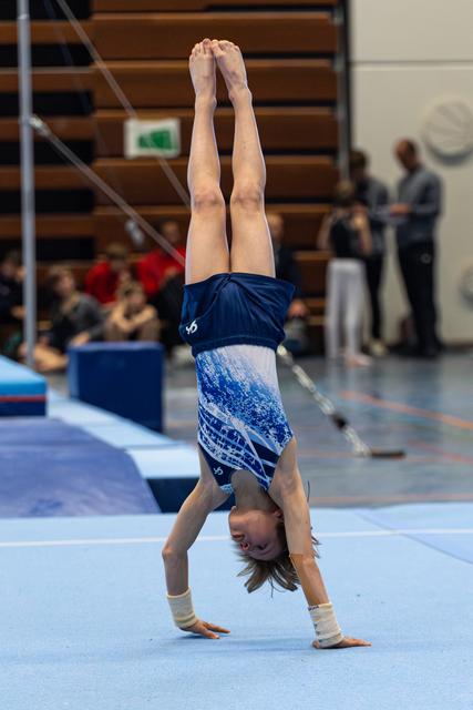Young athlete performs a vertical handstand during floor exercise, wearing blue leotard with white support wraps on wrists