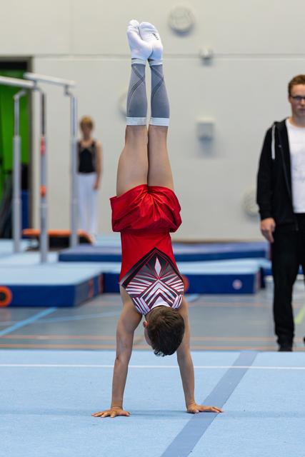 Young athlete performs a handstand on the floor mat, wearing a red leotard and blue shorts, with coaches observing