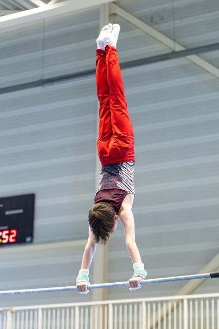 Young athlete performing a handstand on the horizontal bar, wearing a red jacket and patterned leggings in an indoor facility