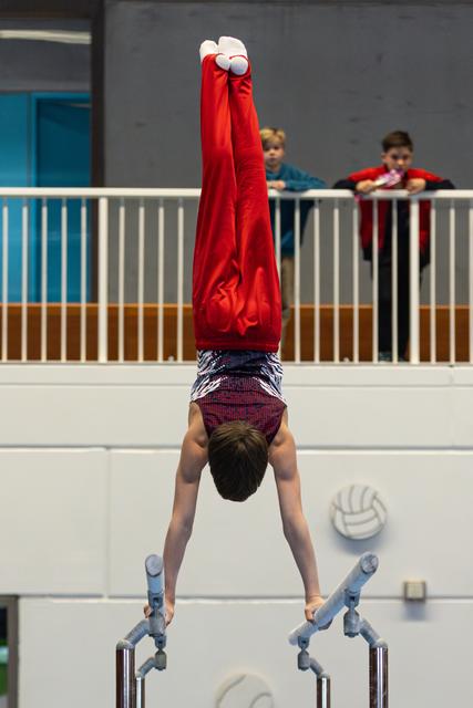 Young athlete performing a handstand on parallel bars with pointed toes, wearing red shirt, while spectators watch from balcony