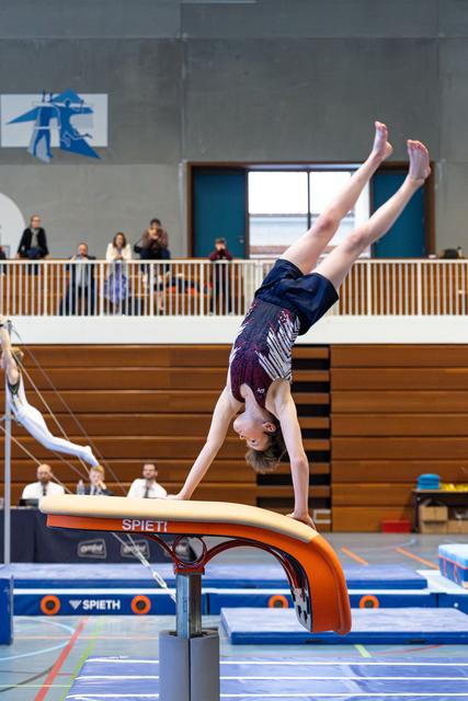 Young female gymnast performs a handstand on the vault table during a competition, spectators watching from the balcony