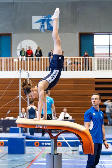 Athlete performs a handstand on vault apparatus while coach spots nearby during indoor sports event