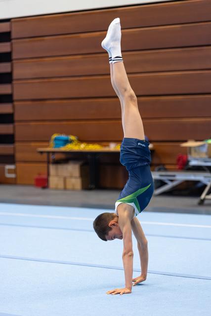 Young athlete performs a perfect handstand with straight legs on a blue gymnastics floor mat in an indoor training facility