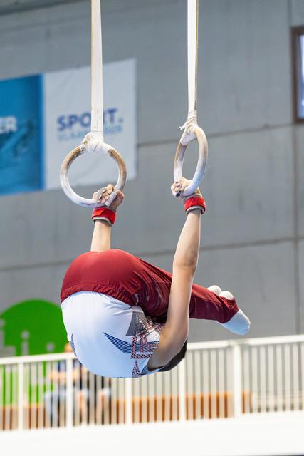 Athlete performing an inverted hang on still rings with legs tucked, wearing red and white uniform at indoor sports facility
