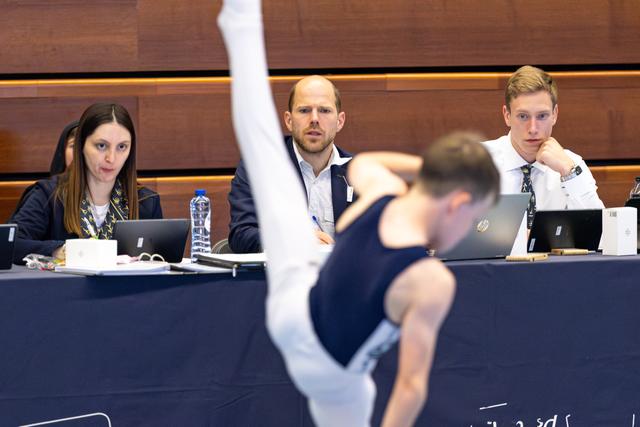 Three judges at table with laptops observe athlete mid-routine with focused expressions during evaluation