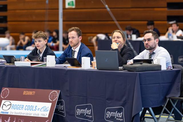 Four judges seated at a table with laptops, evaluating performances during a gymnastics competition in an indoor sports hall