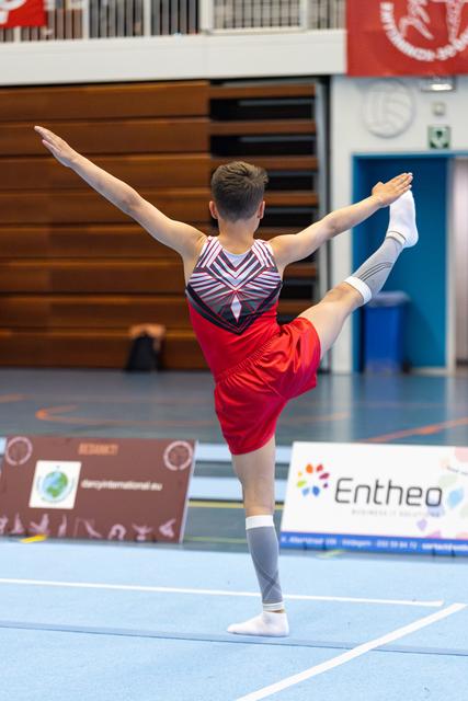 Male athlete performs graceful arabesque pose during floor routine, arms extended, wearing red and striped leotard