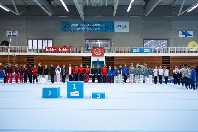 Teams lined up behind medal podium at indoor sports hall, athletes wearing coordinated team tracksuits in red, black, blue, and grey