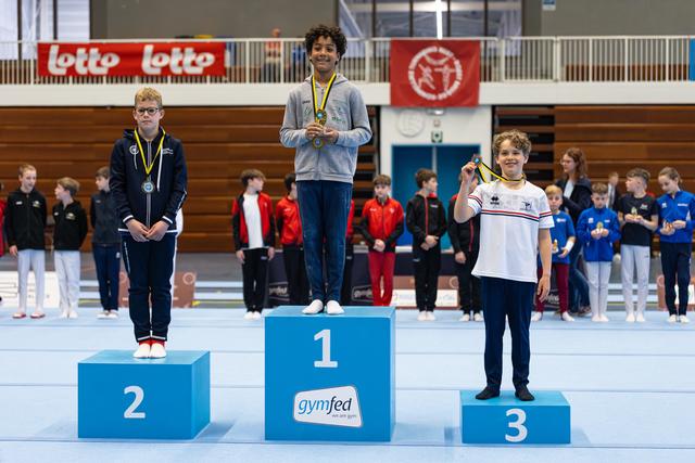 Three young athletes stand on podium with medals, first place celebrating victory while second and third place finishers look on