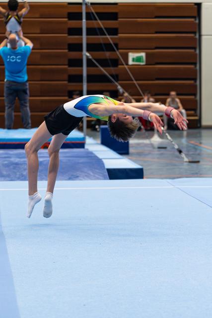 Young athlete executes a dramatic backbend mid-flight during floor exercise, arms extended backwards in indoor training facility