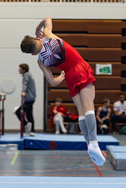 Athlete performs dynamic backbend jump in red uniform and patterned leotard during floor routine at indoor competition