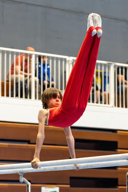 Young athlete performs impressive handstand on parallel bars, legs pointed upward in red attire, spectators watching from balcony