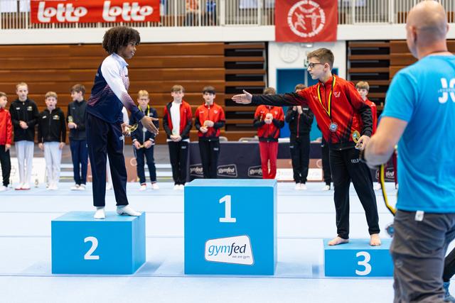 Three young athletes on podium, first place competitor in red pointing at second place winner during medal ceremony