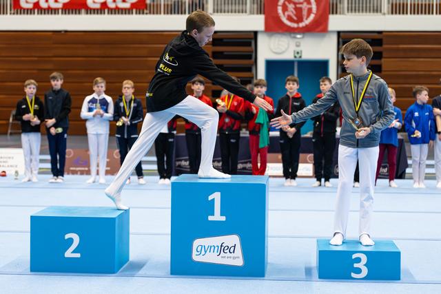 Winner on first-place podium celebrates with high kick while silver and bronze medalists stand beside him in gymnasium