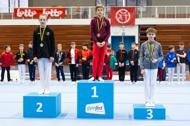Three medalists stand on podium displaying their medals, with first place in burgundy, second in black, and third in gray