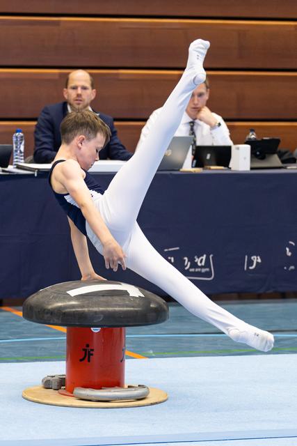 Young athlete performs pommel horse routine with impressive vertical leg extension while judges observe in background