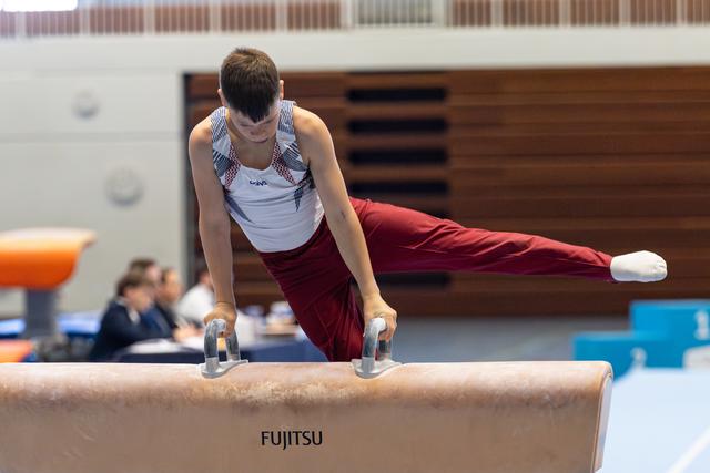 Gymnast performs pommel horse routine with extended legs, wearing maroon pants and white tank top in competition hall