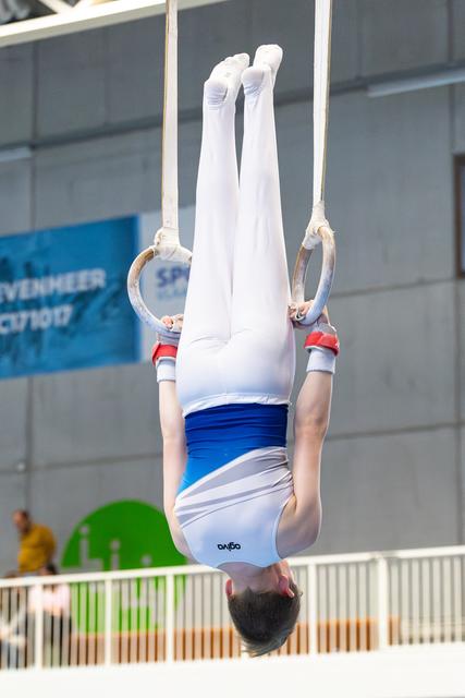 Athlete performing inverted hang on rings with straight body position, wearing white and blue uniform at indoor venue
