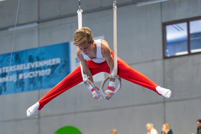 Young athlete performs straddle position on rings, legs extended horizontally, wearing red and white uniform at indoor event