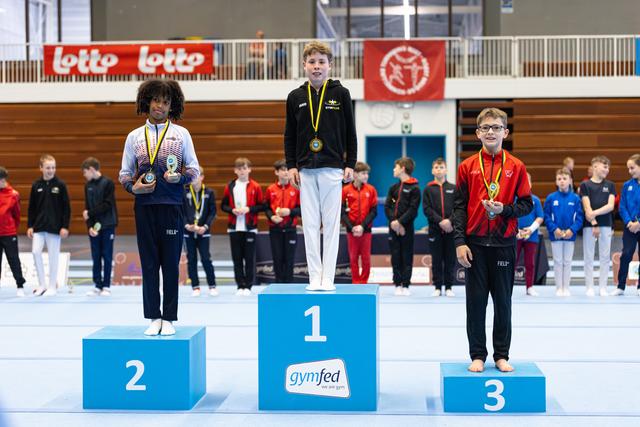 Three medal winners stand on podium positions 1-2-3, wearing their awards with proud expressions at an indoor sports venue