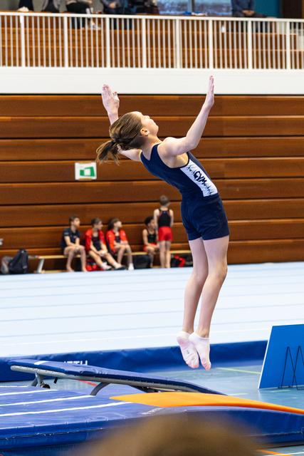 Female trampolinist performs an elegant backbend pose on trampoline, arms raised gracefully overhead in indoor sports hall