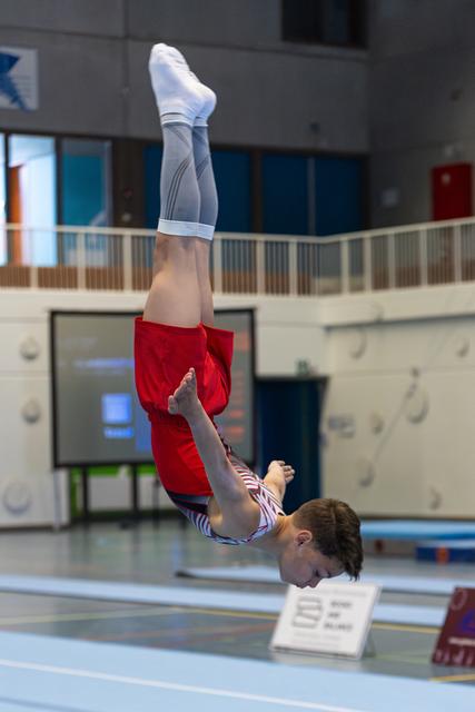 Athlete performs inverted position during floor routine, body fully extended in red and white striped uniform at indoor sports venue