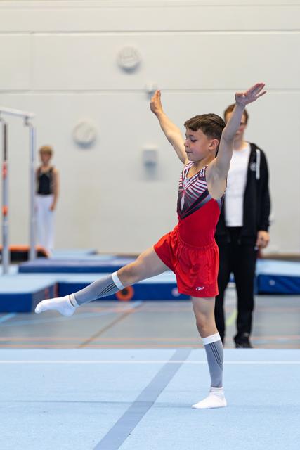 Young athlete performs arabesque on floor exercise, wearing red shorts and grey knee socks, with coach observing in background