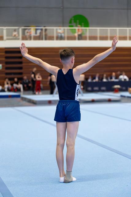 Young athlete stands with arms raised in victorious pose on blue floor mat, facing away from camera during indoor event