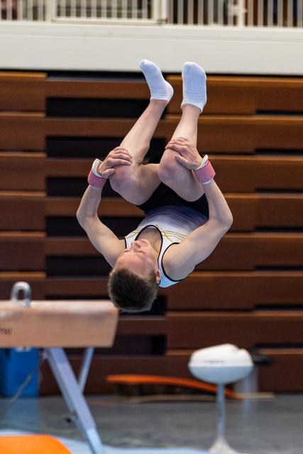 Young athlete performing a backflip in midair during training, gripping legs in tucked position above gymnasium equipment