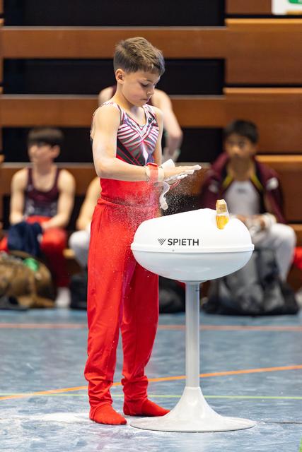Young athlete in red outfit applying chalk to hands at Spieth apparatus before routine, with teammates seated in background