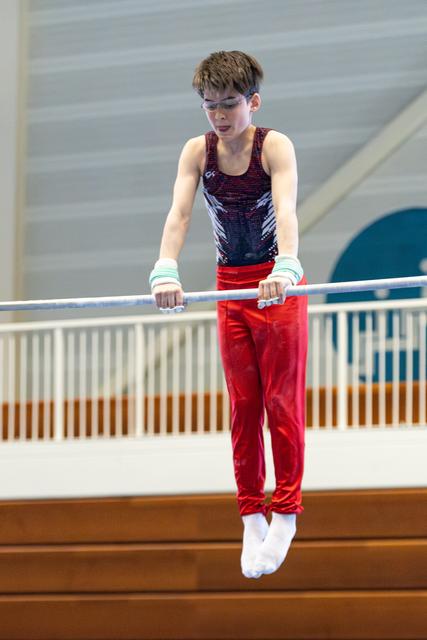Young athlete in red pants and patterned leotard concentrates while performing on horizontal bar with white wrist guards