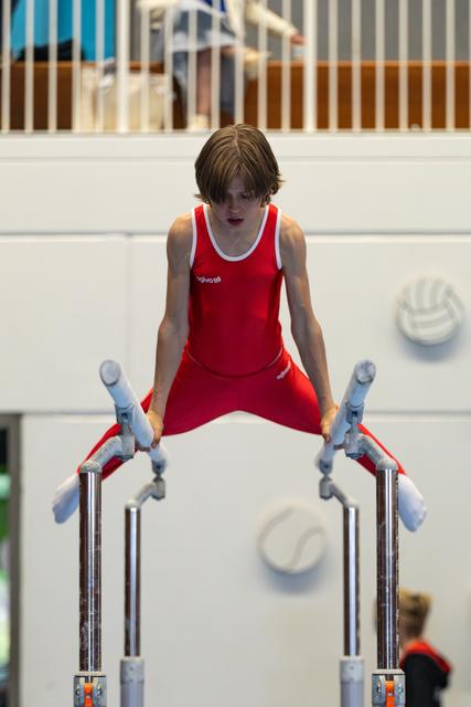 Young athlete in red uniform concentrates intensely while performing on parallel bars at indoor gymnastics event