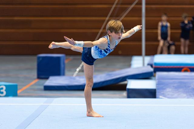 Young gymnast holds a graceful arabesque balance on the floor mat, arms extended with focused concentration during practice