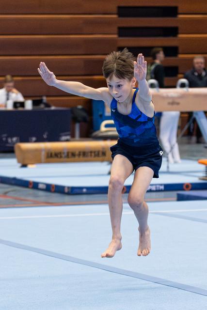 Young gymnast mid-leap during floor routine with arms raised and focused expression at indoor sports venue