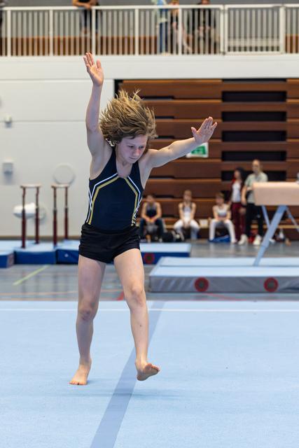 Young athlete landing a tumbling pass on floor exercise, arms raised, hair flowing mid-motion during indoor sports event