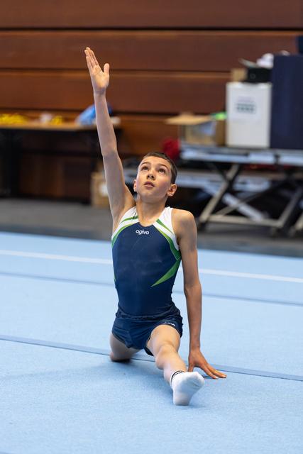 Young gymnast in navy leotard performs elegant finishing pose on floor, one arm raised gracefully while kneeling
