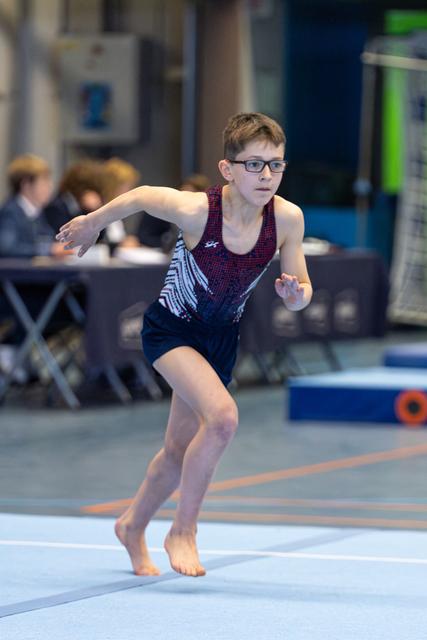 Young athlete in patterned leotard performs floor routine with focused expression and dynamic arm movement during indoor event