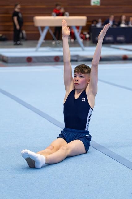 Young athlete performs floor routine with arms raised overhead while seated on the mat, displaying focused expression