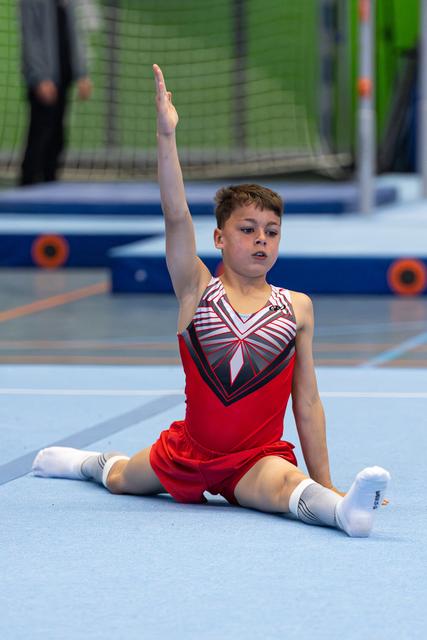 Young gymnast performs floor routine in seated split position with raised arm, wearing red and striped leotard