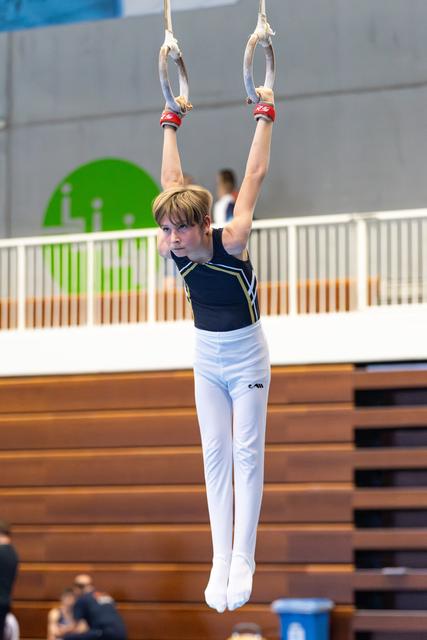 Young athlete performing rings routine with arms extended overhead, showing intense focus and concentration in gymnasium
