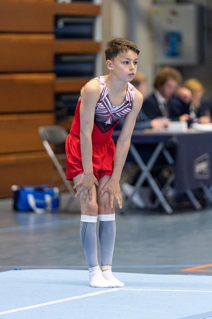 Young athlete in red shorts and striped leotard stands ready on floor mat, hands on knees, displaying focused concentration