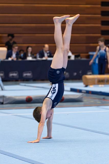 Young athlete performing a handstand on floor exercise mat during indoor competition with judges seated at table in background