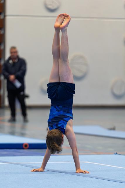Young athlete performing a handstand on blue floor mat with pointed toes, judge visible in background at indoor venue
